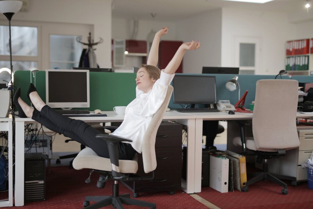 Vista lateral de una alegre empleada vestida formal sentada en una silla de oficina con las piernas cruzadas en el escritorio y estirándose mientras descansa durante el trabajo con los ojos cerrados